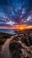 Dramatic sunset over rocky coastal landscape with golden sun descending toward ocean horizon, vibrant orange and blue sky with dynamic clouds, perfect for travel and nature projects.