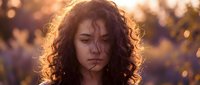 Young woman with curly hair looking down in golden sunset light, purple flowers blurred in background, moody and emotional outdoor portrait.