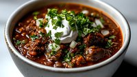 Hearty bowl of spicy chili con carne topped with fresh sour cream, diced onions and chopped parsley garnish in white ceramic bowl, comfort food photography.
