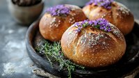 Fresh baked bread rolls decorated with purple edible flowers and herbs on rustic wooden plate, dusted with flour, moody food photography styling.