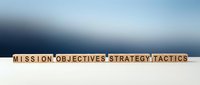 Wooden blocks spelling Mission Objectives Strategy Tactics on a white surface with a blue gradient background, representing business planning concepts.