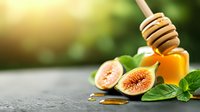 Fresh figs cut in half with honey dipper, golden dripping honey, and green mint leaves arranged on dark stone surface against blurred garden background.