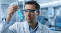Male scientist in safety glasses examining blue liquid sample in test tube during laboratory research experiment.