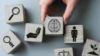 Hand selecting wooden cube with brain icon among business concept blocks including magnifying glass, scales of justice, and human figure symbols on white background.