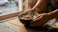 Adult male with medium skin tone holding a ceramic bowl containing fresh salad with mixed greens and vegetables on a wooden table by a window, suitable for food, lifestyle, and culinary content.