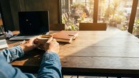 Person working at wooden desk with laptop and coffee cup in bright sunlit home office workspace for remote work and productivity.