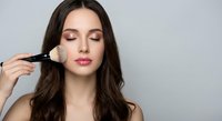 Young brunette woman applying makeup with brush, eyes closed, professional beauty routine for cosmetics and skincare advertising.