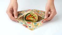 Adult hands holding colorful patterned fabric wrap containing fresh vegetable salad with tomatoes and greens, sustainable food packaging alternative for eco-conscious dining.
