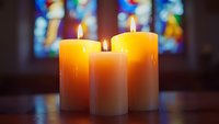Three glowing yellow candles with flames against blurred stained glass windows in church, creating spiritual atmosphere for prayer, worship, or memorial service.