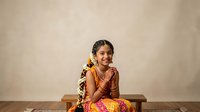 Young South Asian woman wearing traditional red and gold attire with ornamental jewelry, sitting on wooden bench against neutral backdrop for cultural and festive occasions.