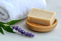 Natural handmade soap bar on wooden dish with lavender flower and green leaves, white towel on marble surface, organic skincare spa concept.
