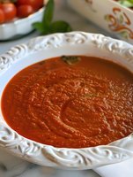 Fresh homemade tomato soup garnished with basil served in ornate white ceramic bowl, close-up view showing smooth, creamy texture and vibrant red color.