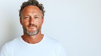 Confident mature Caucasian man with salt and pepper beard wearing white t-shirt against light background, looking at camera with genuine smile. For business or lifestyle design.