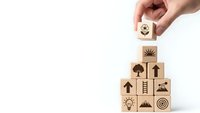Hand placing wooden cube with gear icon on pyramid of blocks featuring business symbols on white background for strategic planning concepts.