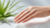 Gentle female hand touching soft white towel in spa setting with green aloe vera plant in background, representing wellness, skincare and relaxation.