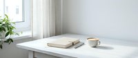 Minimalist workspace with stacked books, ceramic mug, and potted plant on white desk by window with natural light, suitable for lifestyle and office design projects.