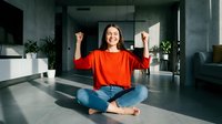 Young Asian woman celebrating success with raised fists while sitting cross-legged on floor in modern living room. Achievement and happiness concept.