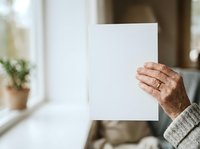 Senior adult male with light skin tone holding blank white paper card indoors near window with potted plant, suitable for mockups, presentations, announcements.