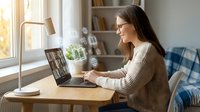 Young woman with glasses working on laptop computer at home office desk near window with natural light and cozy interior decor.