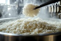 Steaming white rice in metal pot with chopsticks lifting a fluffy portion, kitchen utensils visible in blurred background.