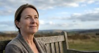 Middle-aged Caucasian woman sitting on wooden bench outdoors enjoying peaceful countryside view with rolling hills and cloudy sky background.