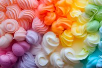 Colorful meringue cookies arranged in rainbow gradient pattern, soft pastel swirls of pink, orange, yellow, green and blue create sweet textured background.