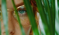 Close up of blue eye with natural freckles peeking through green palm leaves, creating mysterious and romantic mood for beauty and lifestyle content.