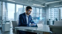 Young male professional, South Asian ethnicity, wearing navy blazer and glasses, working on laptop at modern office desk with city skyline view through floor-to-ceiling windows. Corporate workspace setting.