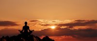 Person meditating in lotus position on rocky mountain peak during golden sunset with dramatic orange sky and clouds for wellness and mindfulness concepts.