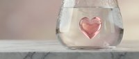 Pink heart-shaped ice cube floating in clear glass of water on marble surface. Romantic drink concept for Valentine's Day celebrations and love themes.
