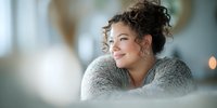 Young mixed-race woman with curly hair smiling warmly in cozy gray sweater, relaxed indoor lifestyle portrait for wellness and happiness concepts.