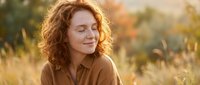 Peaceful redhead woman with curly hair enjoying golden hour sunlight in autumn field, eyes closed in serene moment of mindfulness and relaxation.