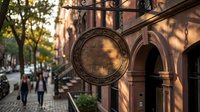 Ornate vintage metal sign hanging from historic building facade with decorative ironwork and warm sunlight creating shadows on cobblestone street.