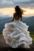 Young woman in flowing white wedding dress with ruffled skirt standing against dramatic sunset sky and mountain landscape, hair blowing in wind, back view.