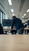 Professional trader analyzing financial charts on computer screen in modern office environment with colleagues working in background.