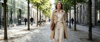 Professional businesswoman in beige trench coat walking confidently on urban street with modern buildings and trees in background during daytime.