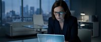 Female professional with dark hair and glasses wearing navy blazer working on laptop in modern high-rise office with city skyline view, suitable for business, corporate, and technology concepts.