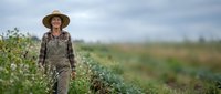 Mature woman farmer wearing straw hat and work clothes standing in agricultural field during daytime, suitable for farming, agriculture, and rural lifestyle content.