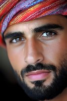 Close-up portrait of Middle Eastern man wearing colorful traditional headscarf with intense gaze and well-groomed beard against dark background.