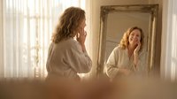 Middle-aged Caucasian woman in white bathrobe applying skincare cream while looking in ornate gold mirror during morning routine in bright bathroom.