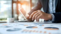Business professional analyzing financial charts and graphs with pen, coffee cup on desk, soft morning light through window, focused on data review and strategy.