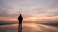 Man standing on beach at sunset gazing at ocean horizon, silhouetted against golden sky and calm water reflections, contemplative mood for travel and lifestyle content.