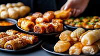 Variety of freshly baked pastries and appetizers on dark plates. Golden brown puff pastry rolls, sesame-topped snacks and savory bites arranged for catering service.