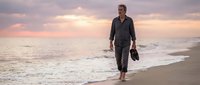 Middle-aged Caucasian male in business attire walking along sandy beach at sunset, carrying shoes, representing work-life balance and career transition concepts.