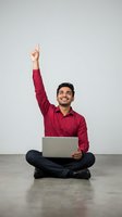 Young South Asian male professional in burgundy shirt and dark pants sitting cross-legged with laptop, raising fist in celebration gesture against white background for success and achievement concepts.