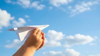 Hand holding white paper airplane against bright blue sky with fluffy white clouds, symbolizing freedom, dreams, and new beginnings in outdoor setting.