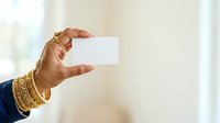 Woman with medium skin tone wearing gold bracelets holding blank white card against blurred neutral background, suitable for business, identity, and promotional mockup uses.