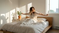 Woman with long curly brown hair stretching on bed in bright bedroom with potted plant and natural sunlight for wellness and lifestyle content.