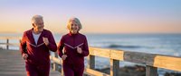 Active senior couple jogging together on wooden boardwalk during golden hour sunset with ocean waves in background for healthy lifestyle and fitness.