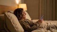 Mature woman with long brown hair relaxing in bed at night, using smartphone while resting against pillows under warm lamp light in cozy bedroom setting.